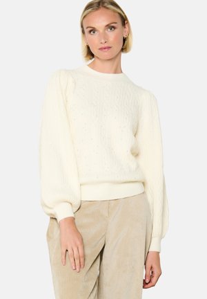 Maglione di filato a cavo crema con maniche a sbuffo, scollo rotondo e dettagli testurizzati. Abbinato a pantaloni in tessuto beige morbido.