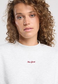 Sweatshirt gris clair avec un col rond et une texture douce. Présente un texte rouge brodé "New York" sur la poitrine. Cheveux bouclés entourant le visage.