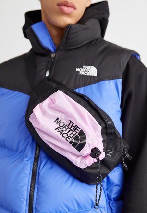 Osoba ubrana w niebiesko-czarną kurtkę The North Face z czarno-różową torbą The North Face przewieszoną przez klatkę piersiową.