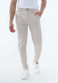 Pantalon tapered beige en mélange de coton, avec deux poches avant, un zip et une seule fermeture à bouton. Assorti avec des baskets blanches.
