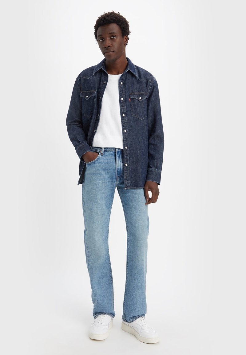 Dunkelblaue Jeanshemd mit Brusttaschen und Knopfverschluss, kombiniert mit hellblauen Jeans und weißen Sneakers. Darunter ein weißes T-Shirt mit Rundhalsausschnitt.
