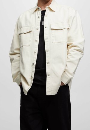 Man wearing an unbuttoned cream shirt jacket over a black t-shirt with black pants, hands in pockets, and a dog tag necklace visible.