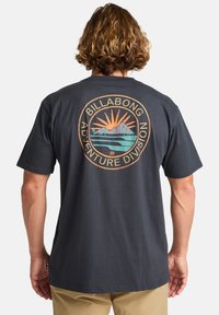 Svart t-skjorte med kort erme som har et sirkulært grafisk design i oransje, grønn og blå på baksiden med teksten "BILLABONG ADVENTURE DIVISION".