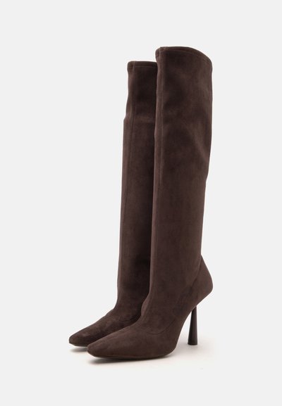 Paire de bottes hautes en daim marron foncé avec bouts pointus et hauts talons aiguilles, debout sur un fond blanc.
