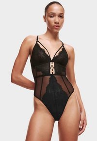 Body en dentelle noire avec panneaux transparents, décolleté en V, bretelles ajustables et un accent géométrique en métal doré à la taille.