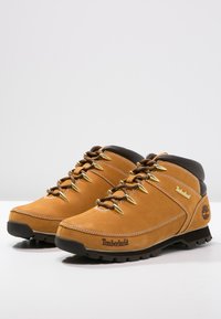 Para beżowych skórzanych butów do wędrówek Timberland z brązowymi wyściółkami, metalowymi oczkami i grubymi czarnymi gumowymi podeszwami na białej powierzchni.