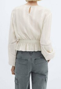 Blusa beige chiaro con maniche lunghe, vita arricciata e orlo a peplum. Presenta una scollatura a chiave sul retro e un motivo testurizzato sottile. Abbinata a pantaloni cargo grigi.