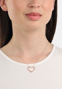 Kvinna som bär ett delikat hjärtformat hänge i roséguld över en vit ribbad t-shirt med rund halsringning, närbild på hals och läppar.