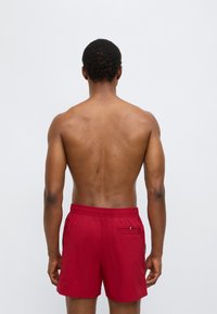 Κόκκινα μαγιό σορτς με ελαστική μέση και πίσω τσέπη. Το ύφασμα φαίνεται ελαφρύ και λείο, κατάλληλο για θαλάσσιες δραστηριότητες.