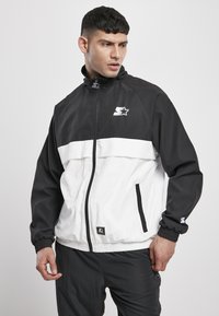 Giacca zip-up in bianco e nero con colletto alto. Presenta polsini elasticizzati, tasche laterali, logo sul petto e un tessuto leggero e texturizzato.