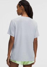 Vrouw met gevlochten haar die een lichtgrijze kortekoope t-shirt en lichtgroene shorts draagt, gezien vanachter tegen een effen achtergrond.