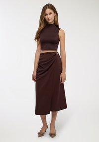 Top corto senza maniche marrone abbinato a una gonna a portafoglio a vita alta con nodo laterale, entrambi realizzati in tessuto liscio. Completano il look le scarpe con tacco marroni.