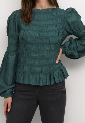 Kvinde iført en grøn bluse med rynkede detaljer og puffede lange ærmer samt sorte jeans, med den ene hånd på hoften foran en ensfarvet baggrund.