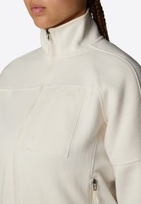 Kobieta nosząca kremowy polar z wysokim kołnierzem i zamkiem błyskawicznym z przodu oraz kieszenią na piersi, z haftowanym logo The North Face na kieszeni.