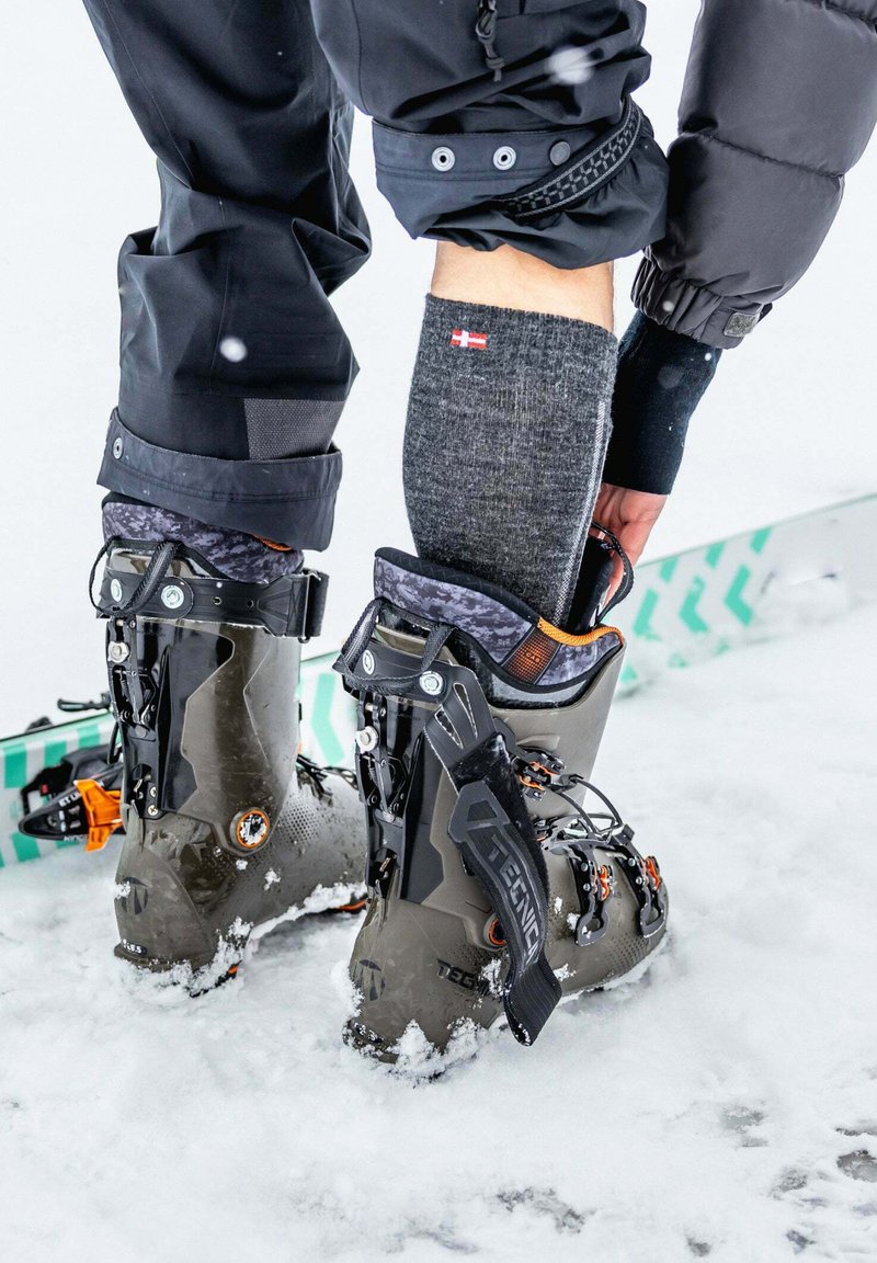 Skischoenen in grijs met zwarte en oranje accenten, een gladde plastic buitenkant, dikke zolen en verstelbare banden, geplaatst in de sneeuw met ski-uitrusting in de buurt.