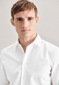 Camicia bianca con colletto a punta, vestibilità sagomata, tessuto liscio e dettagli di cucitura visibili, indossata da un modello su uno sfondo neutro.