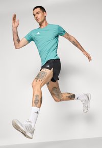 Turquoise athletic t-shirt, black shorts with side slits, white socks, and grey running shoes. Notable tattoos on legs.