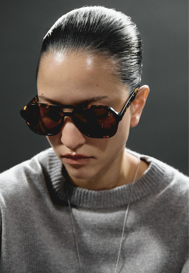 Person with slicked-back hair wearing dark tortoiseshell sunglasses and a gray knit sweater against a dark gray background.