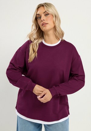Femme blonde portant un sweat à manches longues violet par-dessus un haut blanc et un jean bleu clair, debout devant un fond uni.
