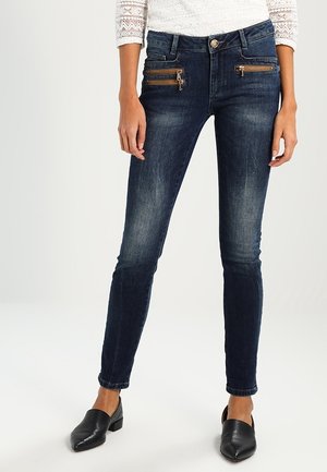 Femme portant un jean skinny bleu foncé avec des poches avant zippées et des chaussures noires à talons bas et bouts pointus.
