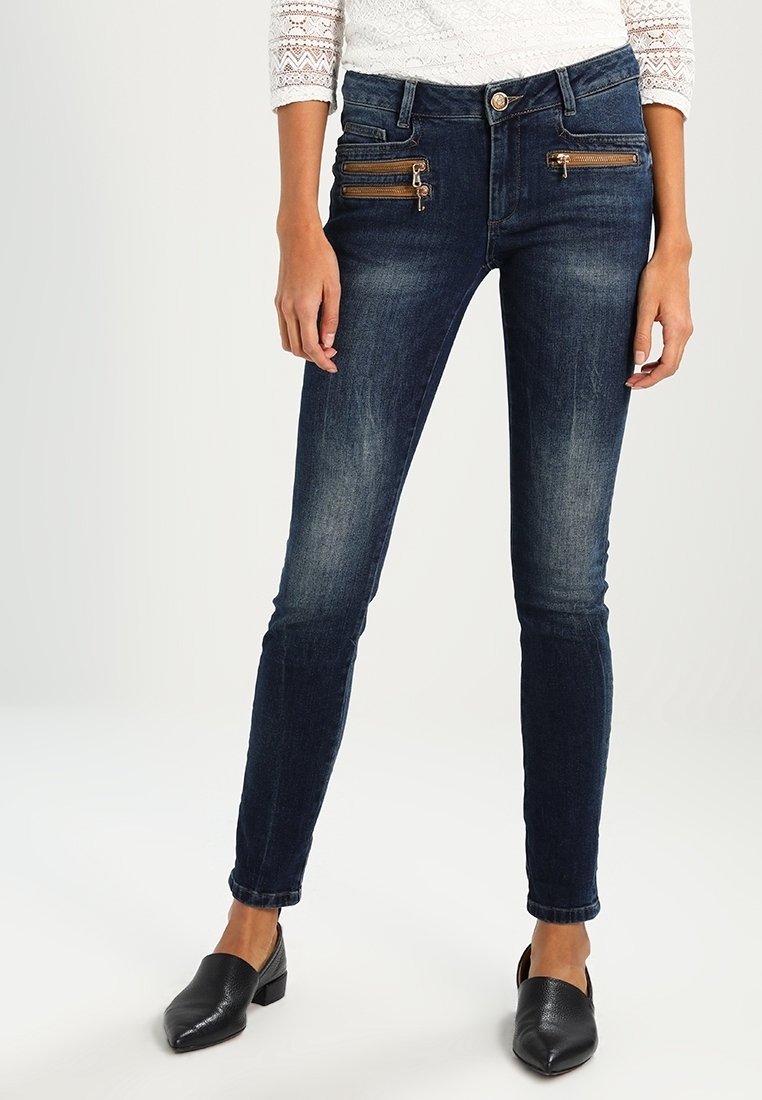 Femme portant un jean skinny bleu foncé avec des poches avant zippées et des chaussures noires à talons bas et bouts pointus.