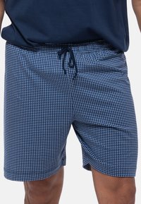 Athlet in marineblauen Badehosen mit einem hellblauen Karomuster. Ein einfarbiges marineblaues Oberteil bedeckt teilweise die Shorts.