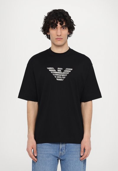 Tricou din bumbac negru, cu guler rotund și croi supradimensionat, având un design cu logo argintiu pe față. Purtați-l cu blugi de culoare albastru deschis.
