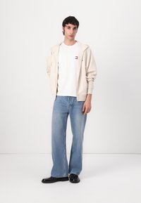 Cremefarvet zip-up hoodie med snørebånd, hvid t-shirt med et lille logo, lyseblå wide-leg jeans og sorte lædersko.