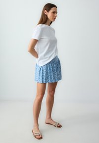 Weißes kurzärmliges Hemd kombiniert mit einem blauen, floral bedruckten Minirock, der eine geraffte Textur und eine lockere Passform aufweist. Tragen mit beigen Riemchensandalen.