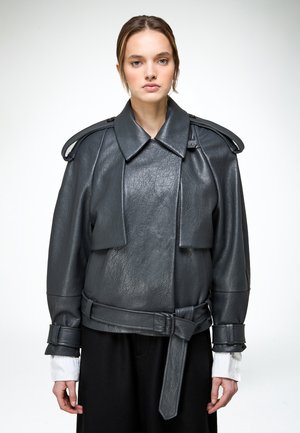 Frau trägt eine strukturierte, schwarze Lederjacke in Übergröße mit weitem Kragen, Schulterklappen, tailliertem Gürtel und sichtbaren weißen Hemdärmeln.