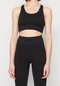 Femme portant un soutien-gorge de sport Puma noir sans coutures et un legging taille haute avec un motif texturé subtil, se tenant devant un fond uni.