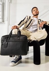 Borsa per laptop in pelle nera con doppio manico, dettagli testurizzati e logo impresso. Indossata con pantaloni chiari e sneakers blu navy.