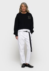 Czarny klasyczny sweter ze haftem "10", w połączeniu z białymi spodniami typu tapered z czarnym paskiem. Model nosi czarne loafersy.