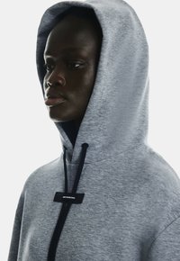 Grauer Kapuzenpullover mit weicher Textur, ausgestattet mit dunklen Kordeln und einem kleinen Logo-Patch. Die Kapuze ist größer und umrahmt das Gesicht.