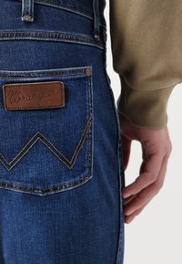 Modre jeans overallke z rjavim usnjenim oznako Wrangler in kontrastnim rumenim šivom. Imajo zadnji žep z zigzag obliko.