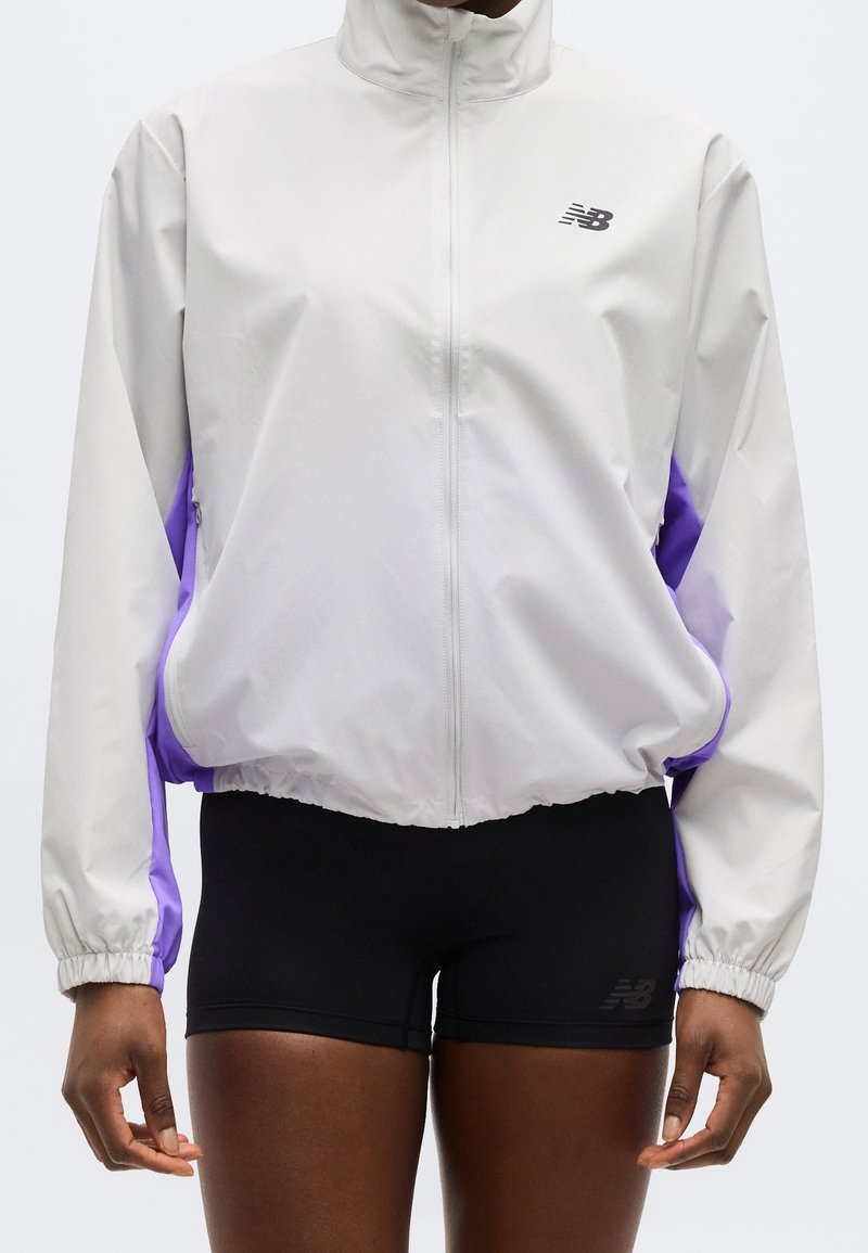 Personne portant une veste blanche zippée avec des panneaux latéraux violets et un short de sport noir, debout devant un fond uni.