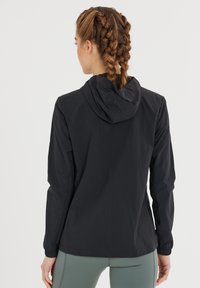 Kvinde med fletninger iført en sort hættetrøje og lysegrå leggings, vent mod en ensfarvet hvid baggrund.