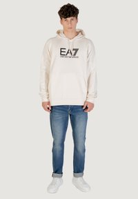 Hoodie in lichtcrème met zwart logo op de voorkant, gecombineerd met blauwe spijkerbroeken en witte sneakers. Zacht materiaal en casual pasvorm.