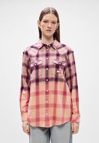 Camisa de flanela xadrez em tons de rosa, roxo e creme, com fecho de botões, dois bolsos no peito e uma bainha curva.