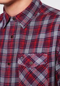 Camicia a quadri rossa e blu navy con colletto button-down, dotata di taschino e bottoni marroni. Il tessuto ha una finitura morbida e testurizzata.