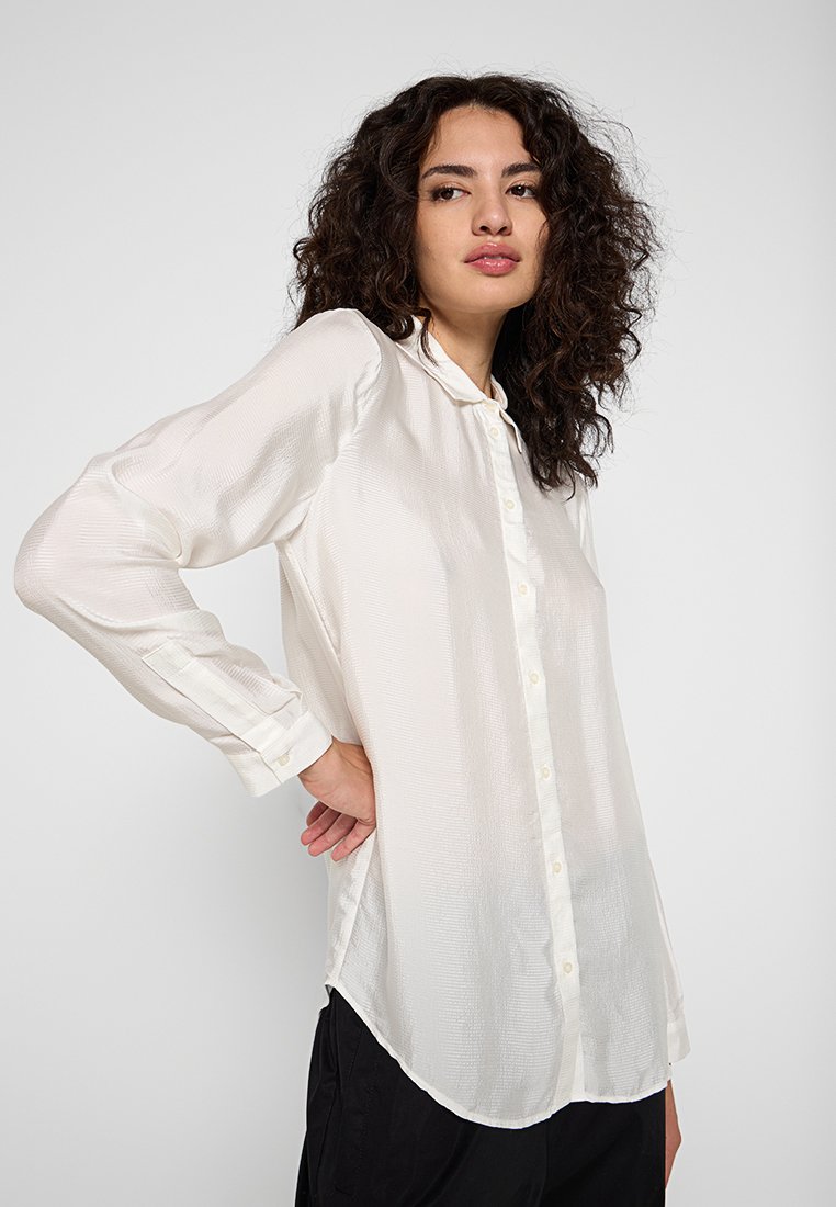 Camisa blanca de manga larga en tejido ligero y texturizado que presenta un frente abotonado y un dobladillo curvado. Diseño con cuello y mangas con puños.