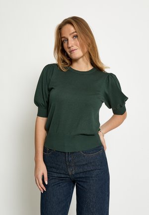 Young woman with light brown hair wearing dark green short-sleeve top and blue jeans, standing against plain light background.
