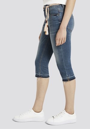 Frau trägt mittelblaue, gekürzte Jeans mit ausgefransten Säumen, einen geflochtenen Gürtel, weiße Turnschuhe und ein weißes Top, seitlich stehend.