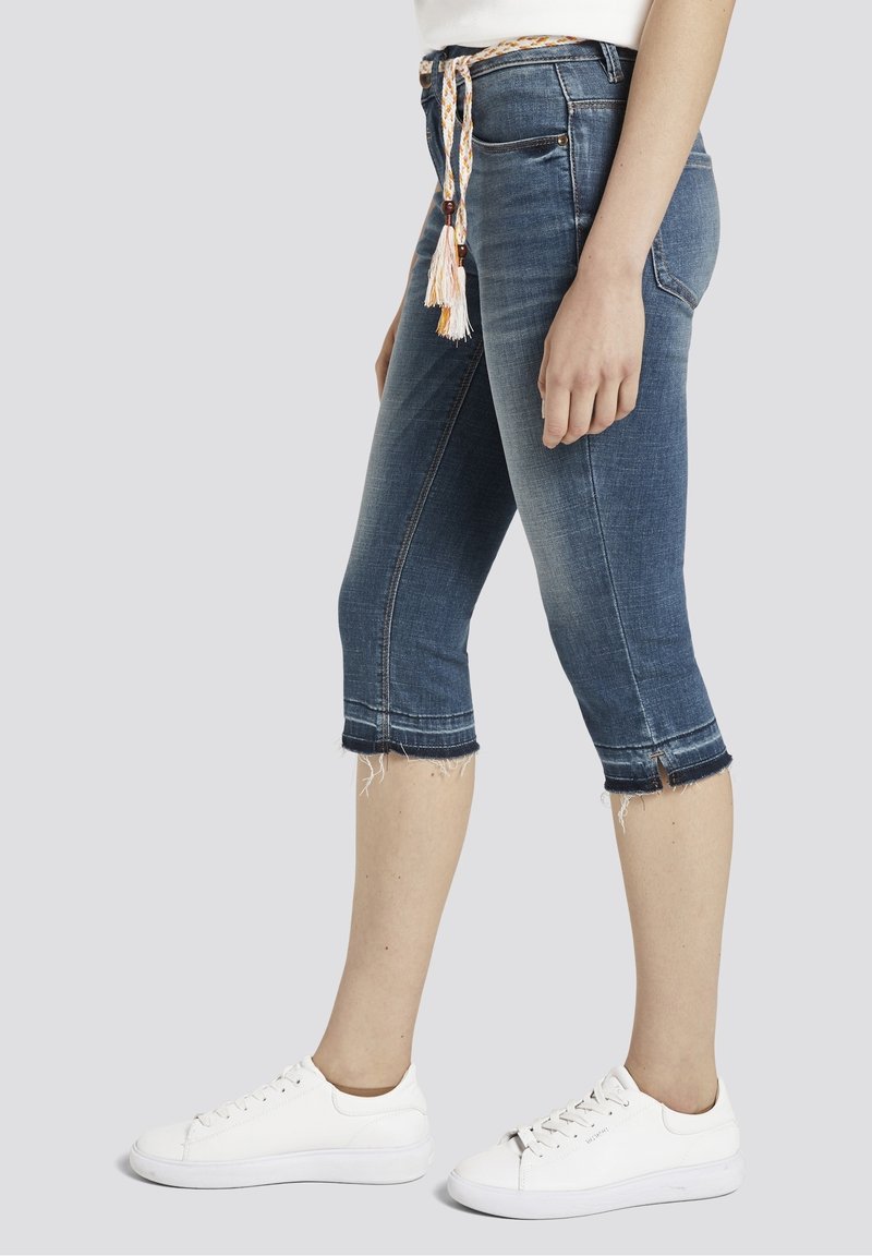 Femme portant un jean denim bleu moyen raccourci avec des ourlets effilochés, une ceinture tressée, des baskets blanches et un top blanc, debout de profil.