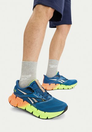 Nohy s šedými ponožkami Reebok, námořnicky modrými šortkami a modrými teniskami Reebok s neonově zelenými a oranžovými podrážkami.
