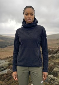 Femme portant une veste polaire noire et un pantalon vert olive, debout en plein air sur un terrain rocheux sous un ciel couvert.