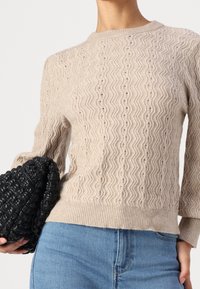 Pull beige en maille avec un motif en zigzag texturé, encolure arrondie et ourlet côtelé, associé à une pochette tissée noire.