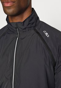 Schwarze Windjacke mit hohem Kragen, mit Reißverschluss, kontrastierenden Nähten und einem kleinen weißen Logo auf der Brust.