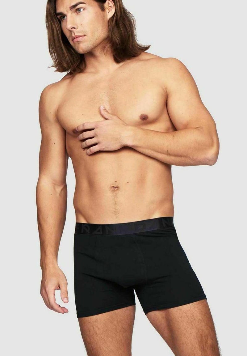 Svarta bomullboxerbriefs med en slät midja och utan synliga mönster. Modellen är poserad något på sidan för att visa passform och täckning.