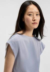 Blusa manica corta azzurro chiaro in un tessuto morbido e fluente. Presenta un colletto a barca e dettagli di cucitura discreti lungo le maniche.